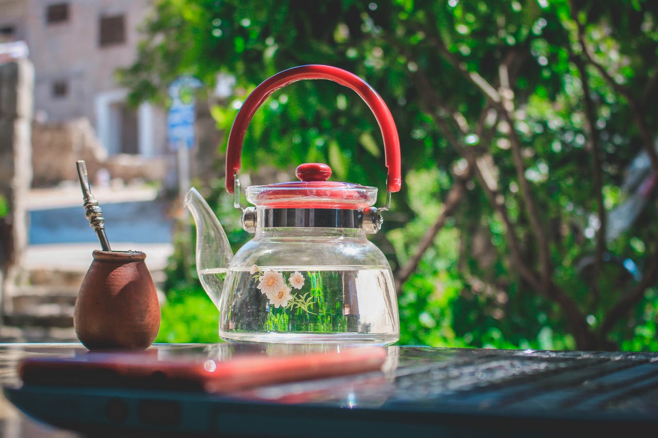 Glass teapot with floral design placed on a wooden table in a sunlit garden, conveying relaxation and summer vibes.